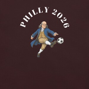 May include: A dark brown t-shirt featuring a graphic of Benjamin Franklin wearing an 18th-century blue coat, tan breeches, and soccer cleats while kicking a black and white soccer ball. The white text above the figure reads PHILLY 2026.