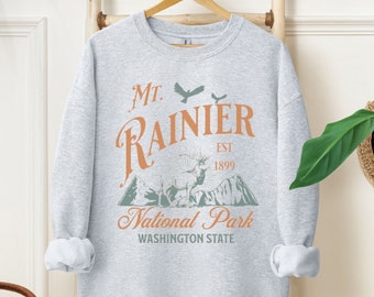 May include: A gray sweatshirt with a vintage-style graphic design featuring the text "Mt. Rainier National Park, Est. 1899, Washington State". The graphic also includes a mountain range, a deer, and a bird.