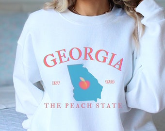 Könnte beinhalten: Weißer Pullover mit einem Pfirsich-Motiv und dem Text "GEORGIA EST 1733 THE PEACH STATE".