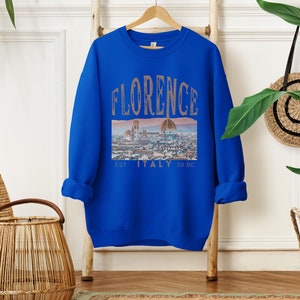 May include: A royal blue sweatshirt with a graphic print of the Florence skyline. The text "FLORENCE EST. ITALY 59 BC" is printed on the sweatshirt.