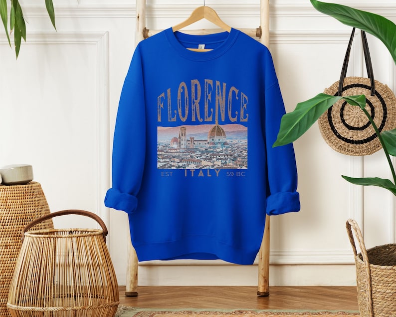 May include: A royal blue sweatshirt with a graphic print of the Florence skyline. The text "FLORENCE EST. ITALY 59 BC" is printed on the sweatshirt.