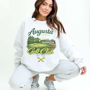 May include: White sweatshirt featuring a colorful graphic of the Augusta Golf Club, with the text "Augusta Georgia Golf Club Est. 1934." The design includes a golf course scene with a flag, trees, and a clubhouse. The sweatshirt is a casual style.