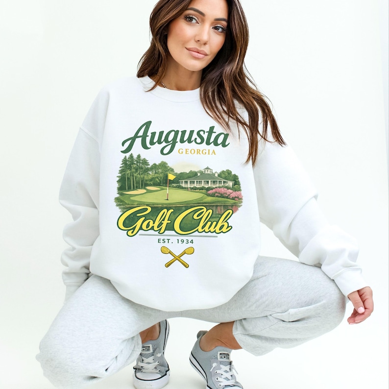 May include: White sweatshirt featuring a colorful graphic of the Augusta Golf Club, with the text "Augusta Georgia Golf Club Est. 1934." The design includes a golf course scene with a flag, trees, and a clubhouse. The sweatshirt is a casual style.
