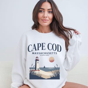 Könnte beinhalten: Weißes Sweatshirt mit Cape Cod-Design. Die Grafik zeigt einen Leuchtturm, den Text "CAPE COD MASSACHUSETTS ATLANTIC COAST", eine Sonne und ein Segelboot. Das Sweatshirt hat einen Rundhalsausschnitt.