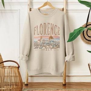 May include: A beige sweatshirt with a vintage-style graphic print of the Florence skyline. The print includes the text "Florence, Est. 59 BC, Italy".