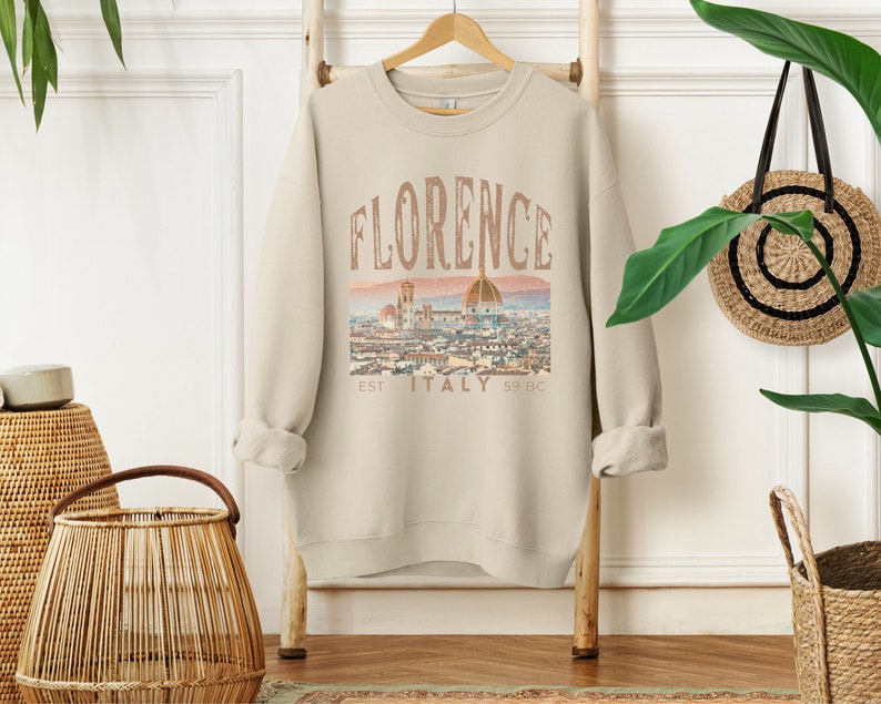 May include: A beige sweatshirt with a vintage-style graphic print of the Florence skyline. The print includes the text "Florence, Est. 59 BC, Italy".