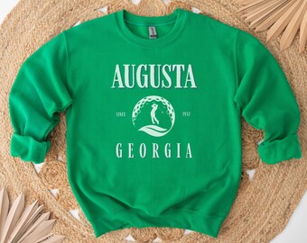 May include: Green sweatshirt with white text that reads "Augusta Since 1932 Georgia" and a white graphic of a golfer in a circle with golf balls.