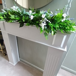May include: A white fireplace mantel with a garland of green leaves and white flowers. The garland is draped over the mantel and hangs down the sides.