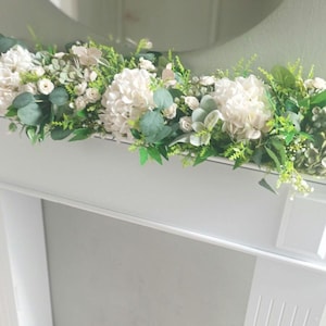 May include: A white fireplace mantel decorated with a garland of white hydrangeas, white roses, and green foliage.