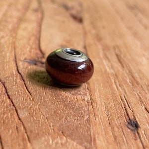 K&ouml;nnte beinhalten: Eine dunkelrote Glasperle mit einem silbernen Band um die Oberseite. Die Perle hat eine gl&auml;nzende Oberfl&auml;che.