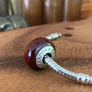 K&ouml;nnte beinhalten: Nahaufnahme eines roten Glasperlenanh&auml;ngers mit silbernen Akzenten. Die Perle ist an einem silbernen Schlangenkettenarmband befestigt. Der Anh&auml;nger tr&auml;gt die Gravur "Pandora".