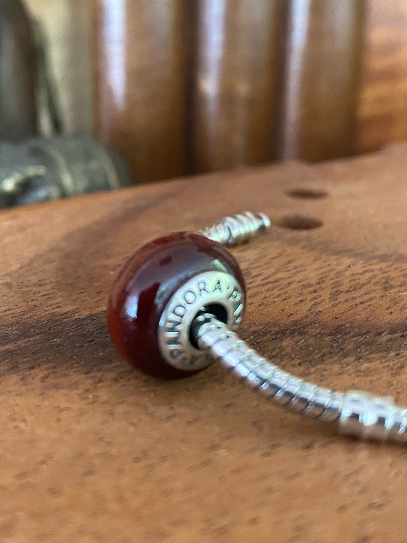 K&ouml;nnte beinhalten: Nahaufnahme eines roten Glasperlenanh&auml;ngers mit silbernen Akzenten. Die Perle ist an einem silbernen Schlangenkettenarmband befestigt. Der Anh&auml;nger tr&auml;gt die Gravur "Pandora".