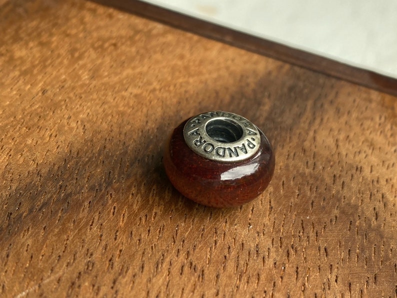 K&ouml;nnte beinhalten: Eine dunkelrote Glasperle mit einem silbernen Metallanh&auml;nger, auf dem "PANDORA" steht.
