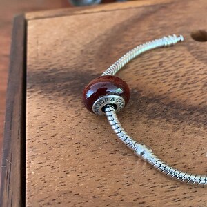 K&ouml;nnte beinhalten: Ein silbernes Schlangenkettenarmband mit einem dunkelroten Glasperlen-Anh&auml;nger, auf dem "Pandora" steht.