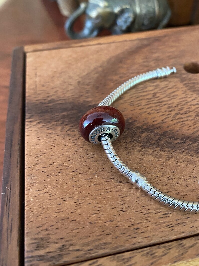 K&ouml;nnte beinhalten: Ein silbernes Schlangenkettenarmband mit einem dunkelroten Glasperlen-Anh&auml;nger, auf dem "Pandora" steht.