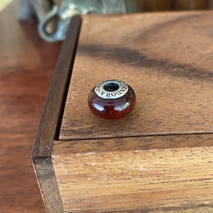 K&ouml;nnte beinhalten: Eine rote Glasperle mit einem silbernen Band, auf dem "PANDORA" steht. Die Perle liegt auf einer Holzoberfl&auml;che.