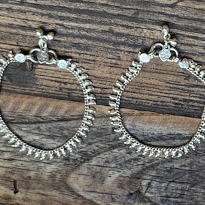 May include: A pair of silver-toned anklets featuring a delicate chain design with small, uniform vertical metal accents along the entire length and a decorative clasp mechanism at the top with tiny dangling charms attached to the circular links.
