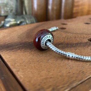 K&ouml;nnte beinhalten: Ein roter Glasperlen-Anh&auml;nger mit der Gravur "PANDORA". Der Anh&auml;nger ist an einem silbernen Schlangenketten-Armband befestigt.