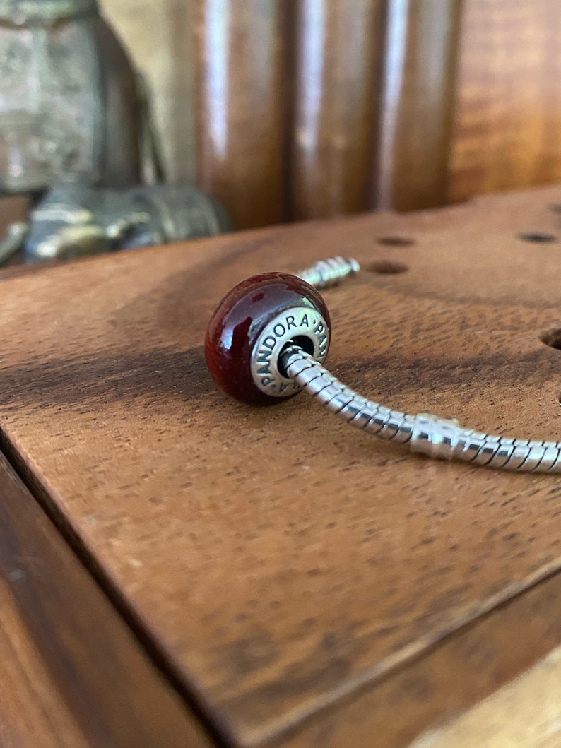 K&ouml;nnte beinhalten: Ein roter Glasperlen-Anh&auml;nger mit der Gravur "PANDORA". Der Anh&auml;nger ist an einem silbernen Schlangenketten-Armband befestigt.