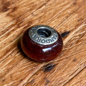 K&ouml;nnte beinhalten: Eine dunkelrote Glasperle mit einem silbernen Metallanh&auml;nger, auf dem "PANDORA" steht.