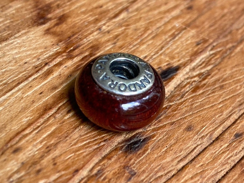 K&ouml;nnte beinhalten: Eine dunkelrote Glasperle mit einem silbernen Metallanh&auml;nger, auf dem "PANDORA" steht.