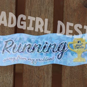 Może przedstawiać: Jasnoniebieski baner z napisem "Running away from my problems" w ozdobnej czcionce. Po prawej stronie znajduje się żółta grafika trofeum z napisem "#1 Most Delulu". Baner umieszczono na drewnianym tle.