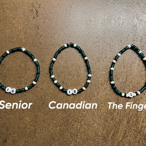 K&ouml;nnte beinhalten: Drei gr&uuml;ne und wei&szlig;e Perlenarmb&auml;nder mit Zahlen darauf. Die Armb&auml;nder sind mit "Senior", "Canadian" und "The Finger" beschriftet.