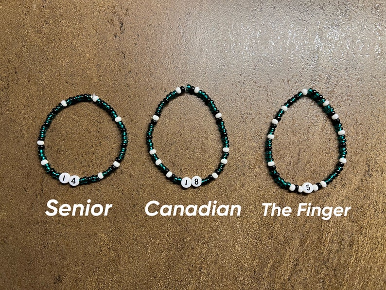 K&ouml;nnte beinhalten: Drei gr&uuml;ne und wei&szlig;e Perlenarmb&auml;nder mit Zahlen darauf. Die Armb&auml;nder sind mit "Senior", "Canadian" und "The Finger" beschriftet.