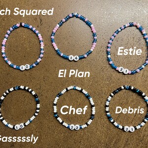 K&ouml;nnte beinhalten: Sechs Perlenarmb&auml;nder mit verschiedenen Farbkombinationen und Namen auf den Anh&auml;ngern. Die Namen sind "French Squared", "El Plan", "Estie", "Gasssssly", "Chef" und "Debris".