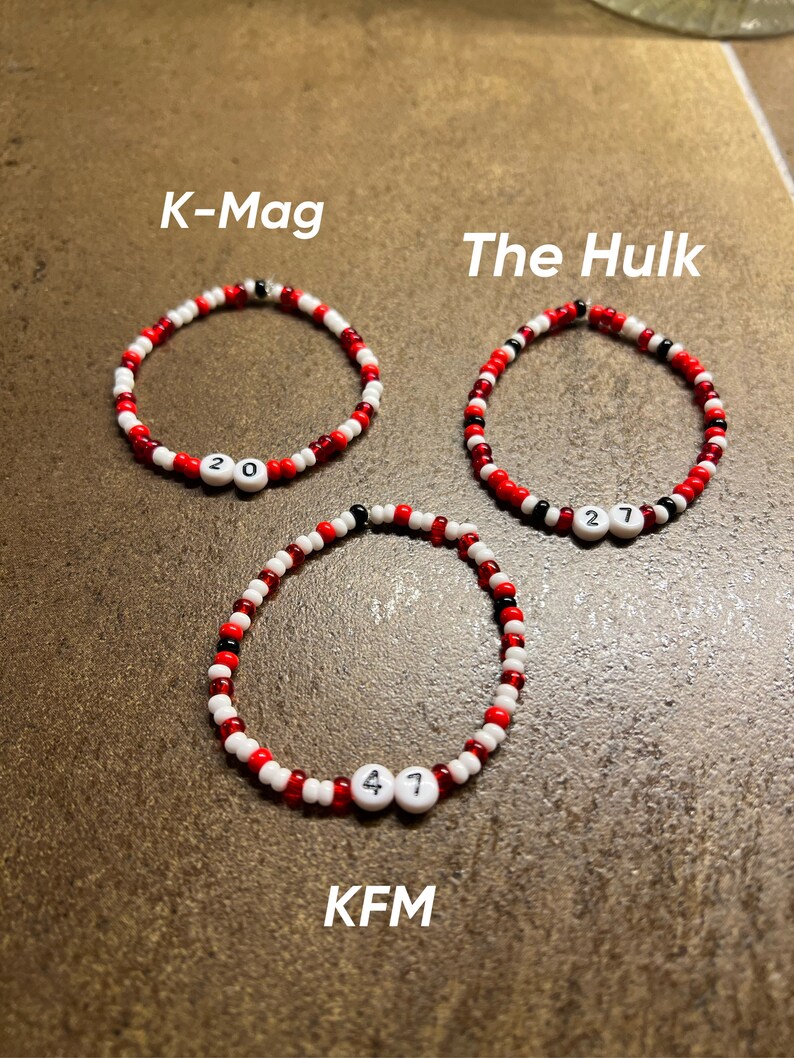 K&ouml;nnte beinhalten: Drei rote, wei&szlig;e und schwarze Perlenarmb&auml;nder mit wei&szlig;en Buchstabenperlen, die "K-Mag 20", "The Hulk 27" und "KFM 47" buchstabieren.