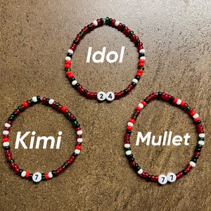K&ouml;nnte beinhalten: Drei rote, wei&szlig;e und gr&uuml;ne Perlenarmb&auml;nder mit den Worten "Idol", "Kimi" und "Mullet" darauf. Jedes Armband hat eine wei&szlig;e Perle mit einer Zahl darauf: 24, 7 und 7.