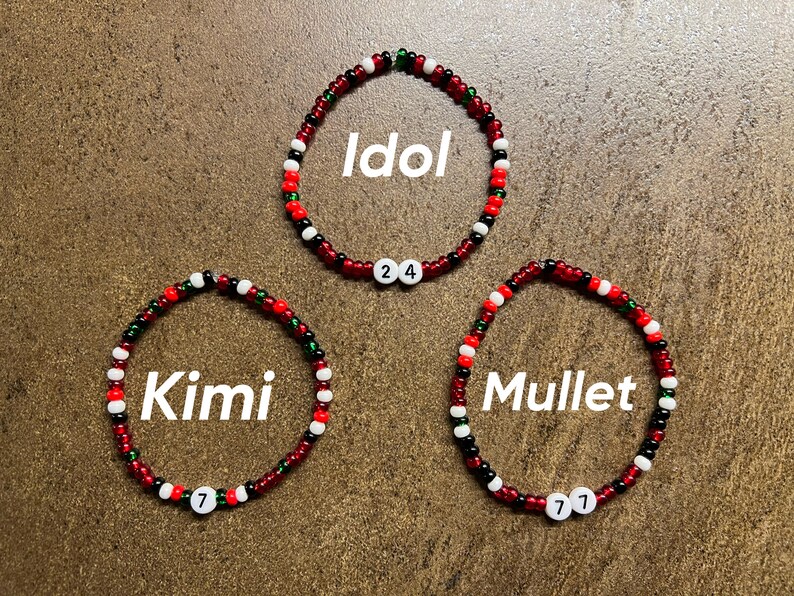 K&ouml;nnte beinhalten: Drei rote, wei&szlig;e und gr&uuml;ne Perlenarmb&auml;nder mit den Worten "Idol", "Kimi" und "Mullet" darauf. Jedes Armband hat eine wei&szlig;e Perle mit einer Zahl darauf: 24, 7 und 7.