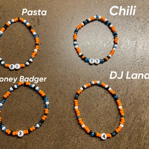 K&ouml;nnte beinhalten: Vier Perlenarmb&auml;nder mit orangefarbenen, schwarzen und wei&szlig;en Perlen. Jedes Armband hat eine wei&szlig;e Perle mit einer Nummer darauf. Die Armb&auml;nder sind mit "Pasta", "Chili", "Honey Badger" und "DJ Lando" beschriftet.