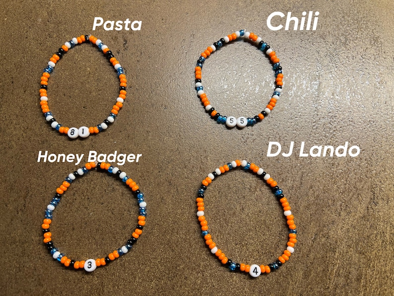 K&ouml;nnte beinhalten: Vier Perlenarmb&auml;nder mit orangefarbenen, schwarzen und wei&szlig;en Perlen. Jedes Armband hat eine wei&szlig;e Perle mit einer Nummer darauf. Die Armb&auml;nder sind mit "Pasta", "Chili", "Honey Badger" und "DJ Lando" beschriftet.