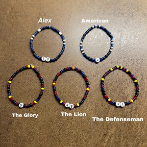 K&ouml;nnte beinhalten: F&uuml;nf Perlenarmb&auml;nder mit verschiedenen Farbkombinationen und Zahlen. Die Armb&auml;nder sind mit "Alex", "American", "The Glory", "The Lion" und "The Defenseman" beschriftet.