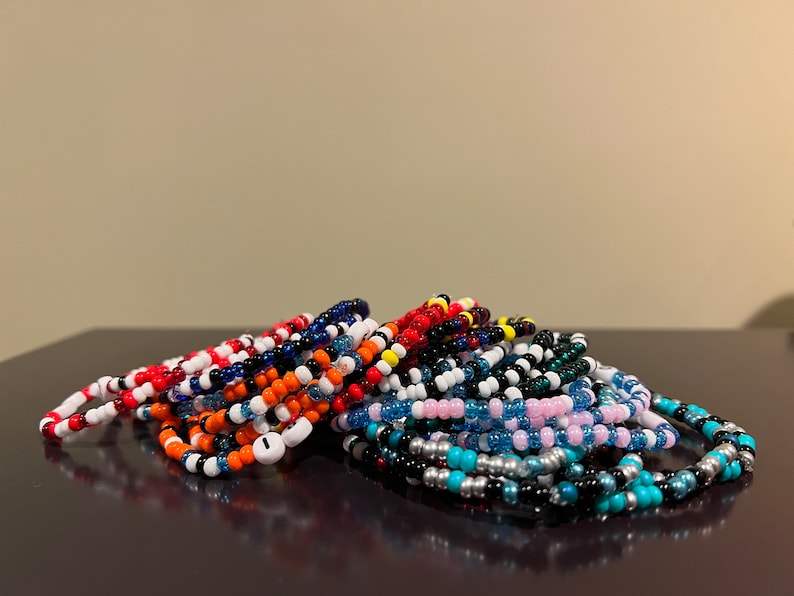 K&ouml;nnte beinhalten: Ein Stapel bunter Perlenarmb&auml;nder in verschiedenen Farben, darunter Rot, Blau, Schwarz, Wei&szlig;, Orange, Pink, T&uuml;rkis und Silber.