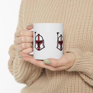 May include: White ceramic mug featuring a red, black, and white helmet design. The design is repeated twice on the mug. The mug is held by a person wearing a beige sweater.