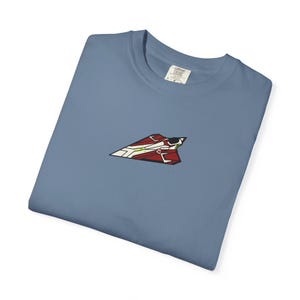May include: A blue t-shirt with a small embroidered design of a red, white, and black airplane. The shirt is made of a soft material and has a classic crew neck.