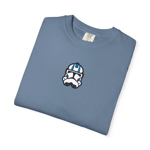 May include: A blue t-shirt featuring a small embroidered image of a white and blue helmet. The helmet has black accents and is centered on the chest of the shirt. The shirt is a solid color and appears to be made of a soft material.