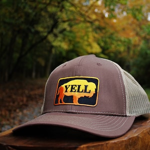 May include: A brown and beige trucker hat with a black and yellow patch featuring a buffalo silhouette and the word "YELL".