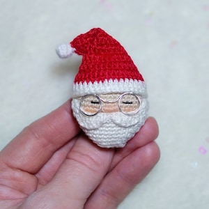 May include: A crocheted Santa Claus head with a red hat, white beard, and silver wire-rimmed glasses. The Santa Claus head is a festive decoration, perfect for the holiday season. The hat has a white pom-pom.