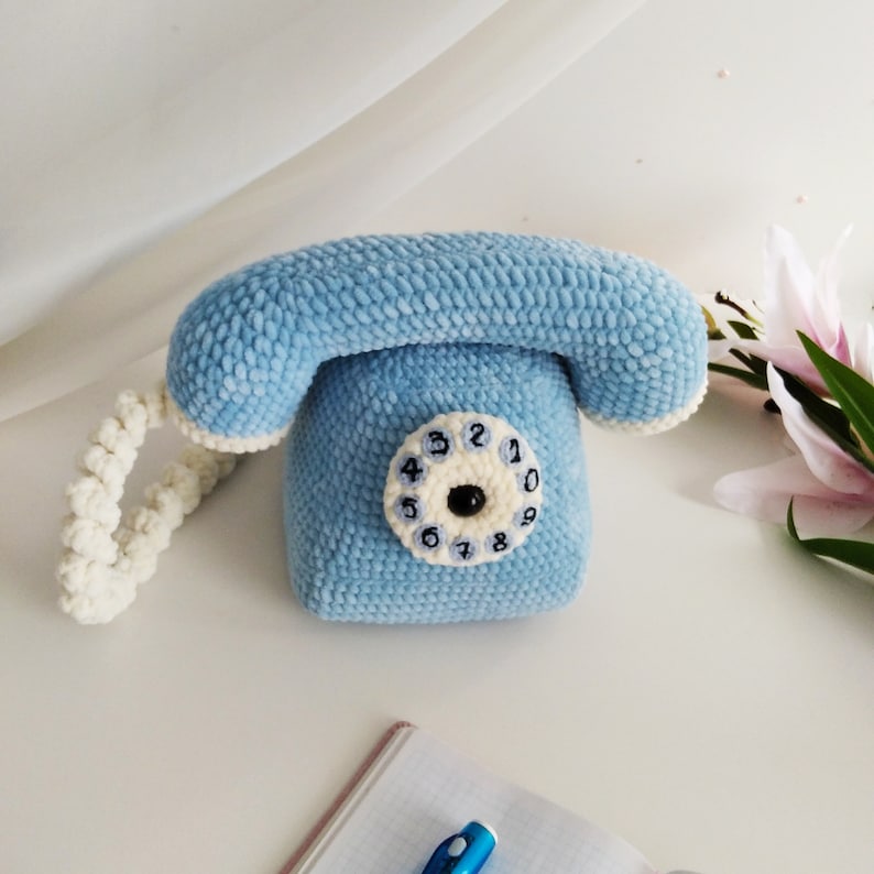 May include: A blue crocheted rotary phone with a white cord and a white dial with black numbers. The phone is sitting on a white surface.