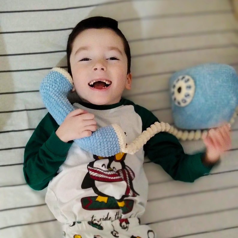 May include: A young child in a white and green shirt with a penguin design is lying on a striped bedspread. The child is holding a blue and white crocheted toy phone to their ear and smiling.