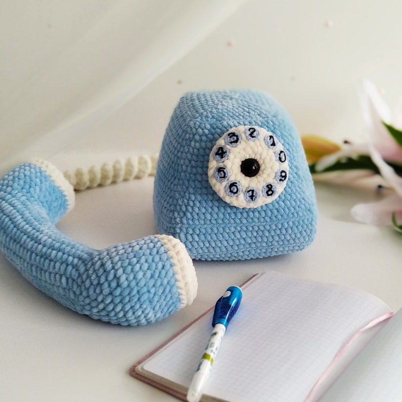 May include: A blue crocheted toy telephone with a white dial and black numbers. The phone has a receiver and a base. The phone is sitting on a white surface.