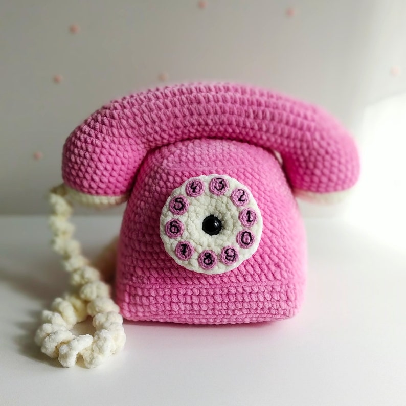 May include: A pink crocheted toy telephone with a white dial and black numbers. The phone has a white cord.