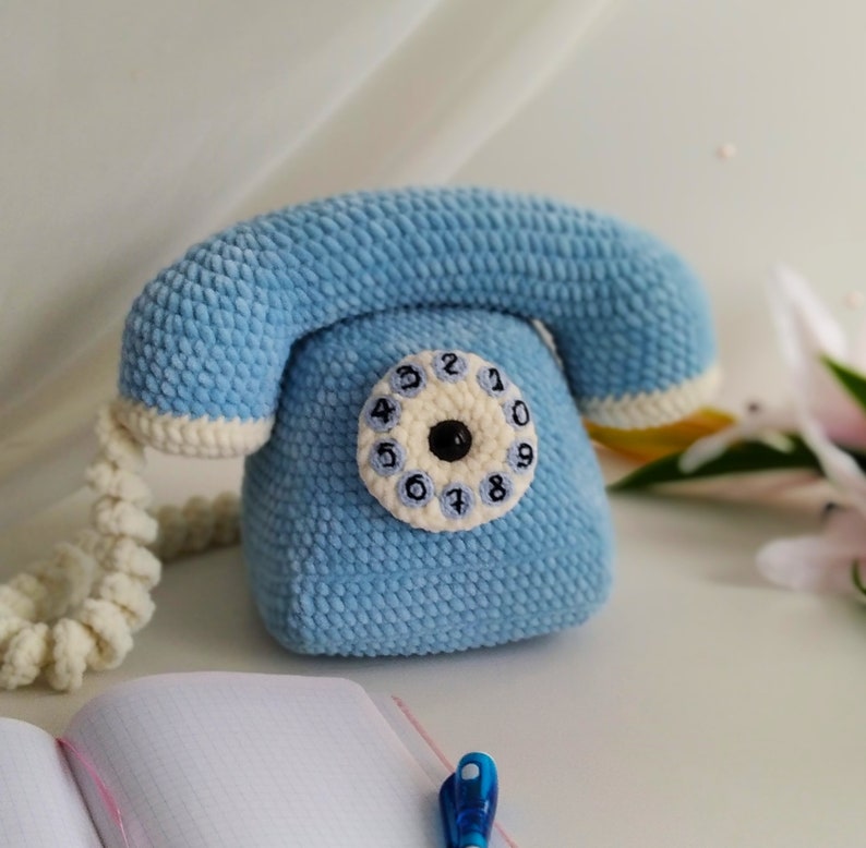 May include: A blue crocheted toy telephone with a white dial and black numbers. The phone has a white cord.