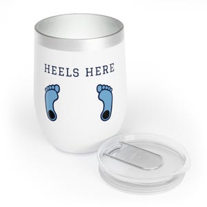 May include: White stainless steel tumbler with a silver lid. The tumbler has a blue and white footprint design and the text "HEELS HERE" printed on the side.