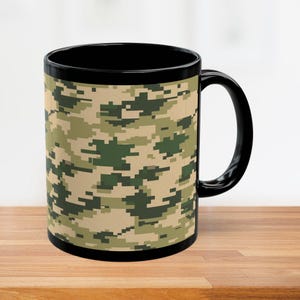 May include: Black ceramic mug with a camouflage pattern in shades of green, tan, and brown. The mug has a black handle and rim, and it sits on a wooden surface.