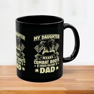 May include: Black ceramic mug with the text "MY DAUGHTER WEARS COMBAT BOOTS PROUD MILITARY DAD" in white. The design includes combat boots and American flags. The mug is on a wooden surface.