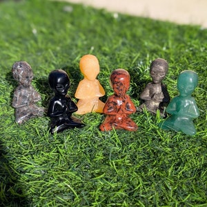 May include: Six small, hand-carved figurines of meditating figures made from different types of stone. The figurines are in various colours, including black, green, brown, grey, and orange.